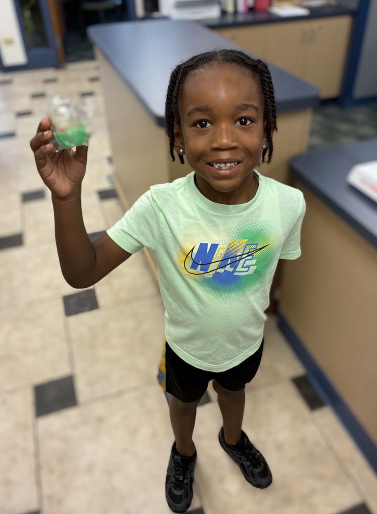 A smiling child wearing a light mint green Nike T-shirt and black shorts, holding up a small wrapped green toy in their right hand.