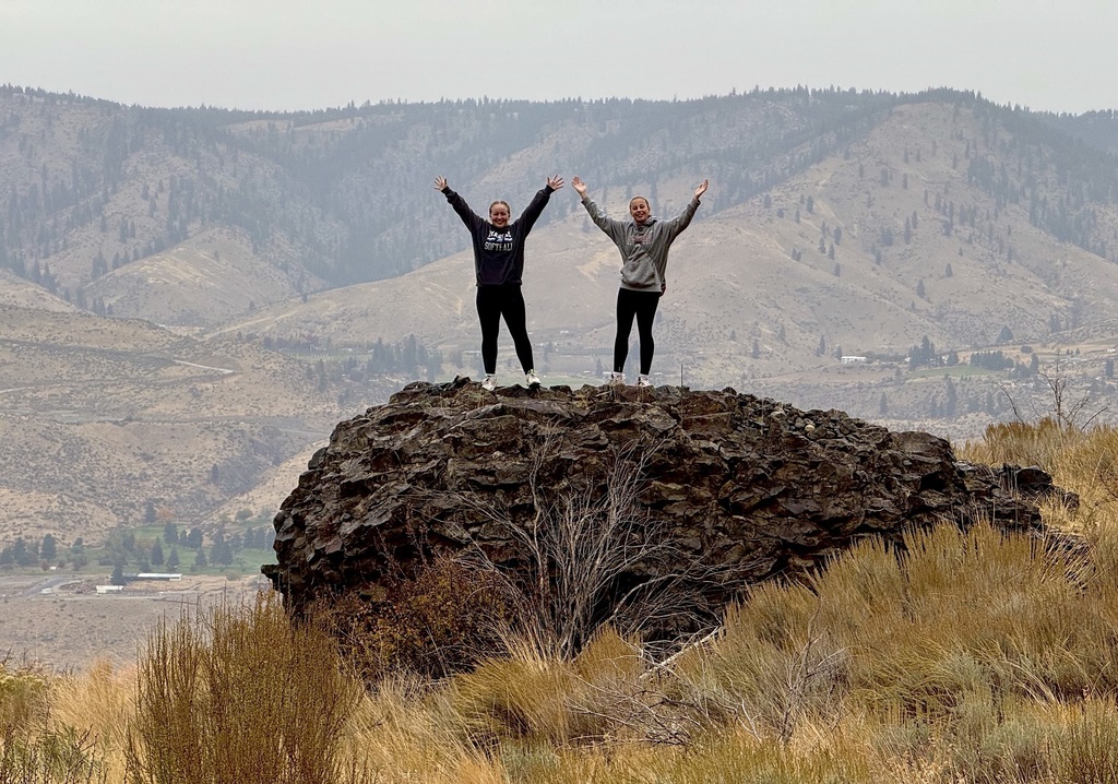 Two female students pose on top of a rock with their arms in the air.