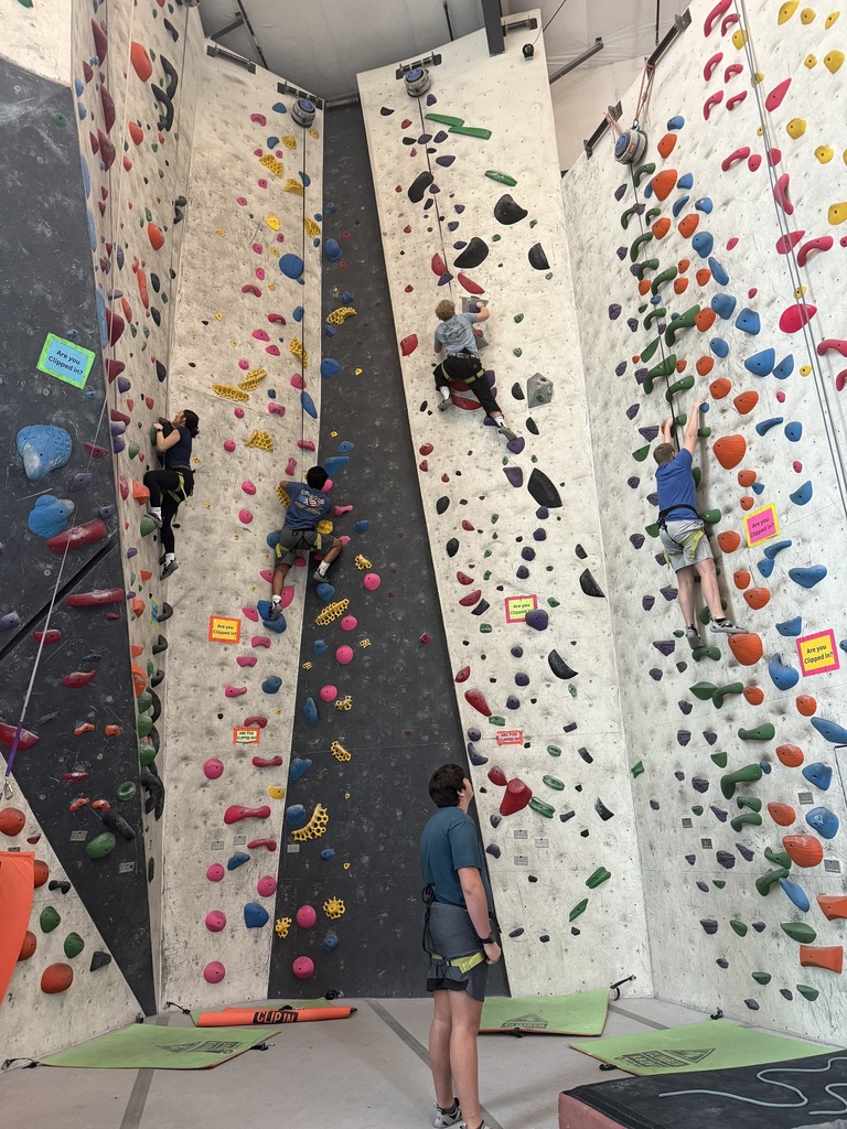 Four students climb a rock wall while a boy looks on.
