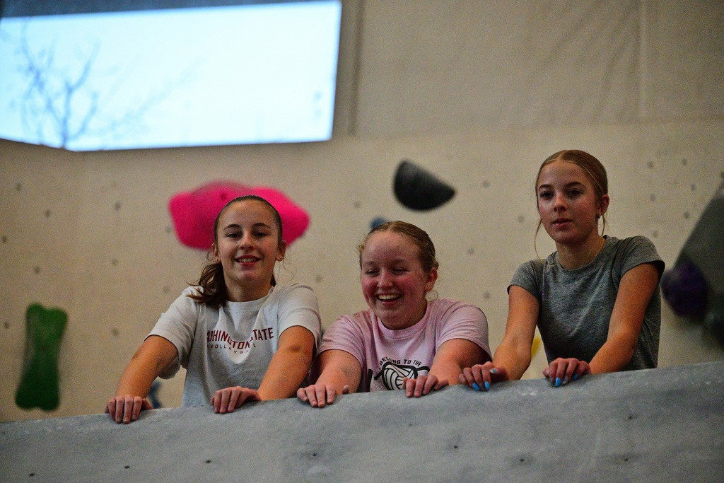 Three girls look down from the top of the climbing wall.