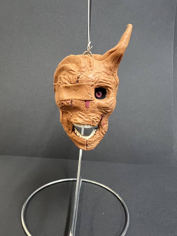 Horror Mask – Front View: A creepy clay monster mask with wrinkles, sharp teeth, and one exposed eye displayed on a stand.