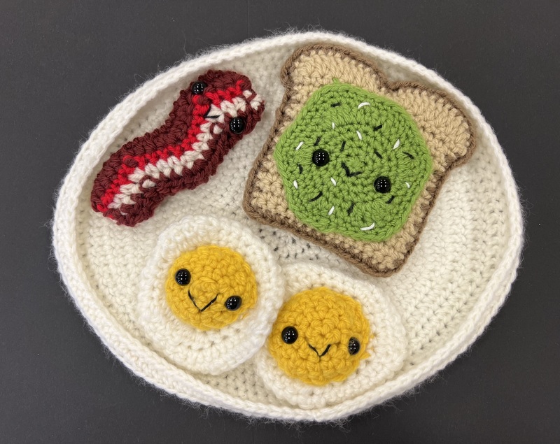 A crocheted breakfast plate featuring smiling eggs, bacon, and avocado toast.