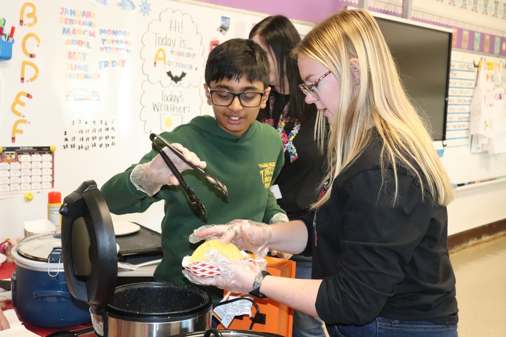 A student uses tongs to place meat on a taco held by a teacher