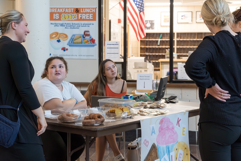  Two students sit at a bake sale table while adults stand nearby smiling and talking with them.