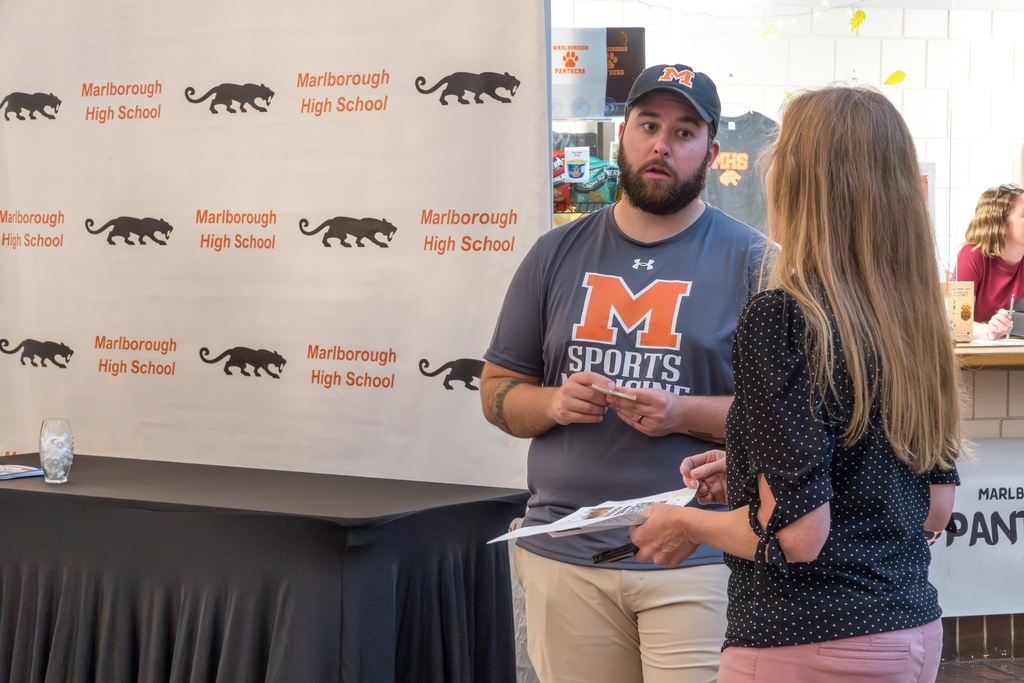 An adult wearing a Marlborough sports t-shirt and hat talks to a woman. A Marlborough High School backdrop is behind them.