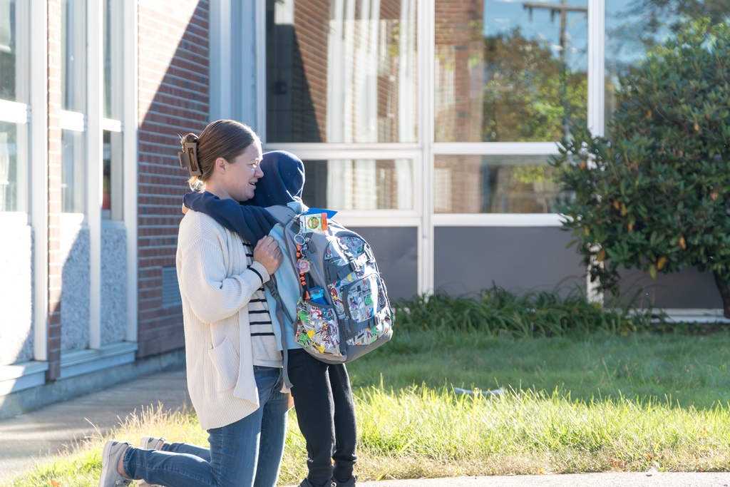 A Richer School mom kneels on the sidewalk, holding onto her son, who stands and hugs her.