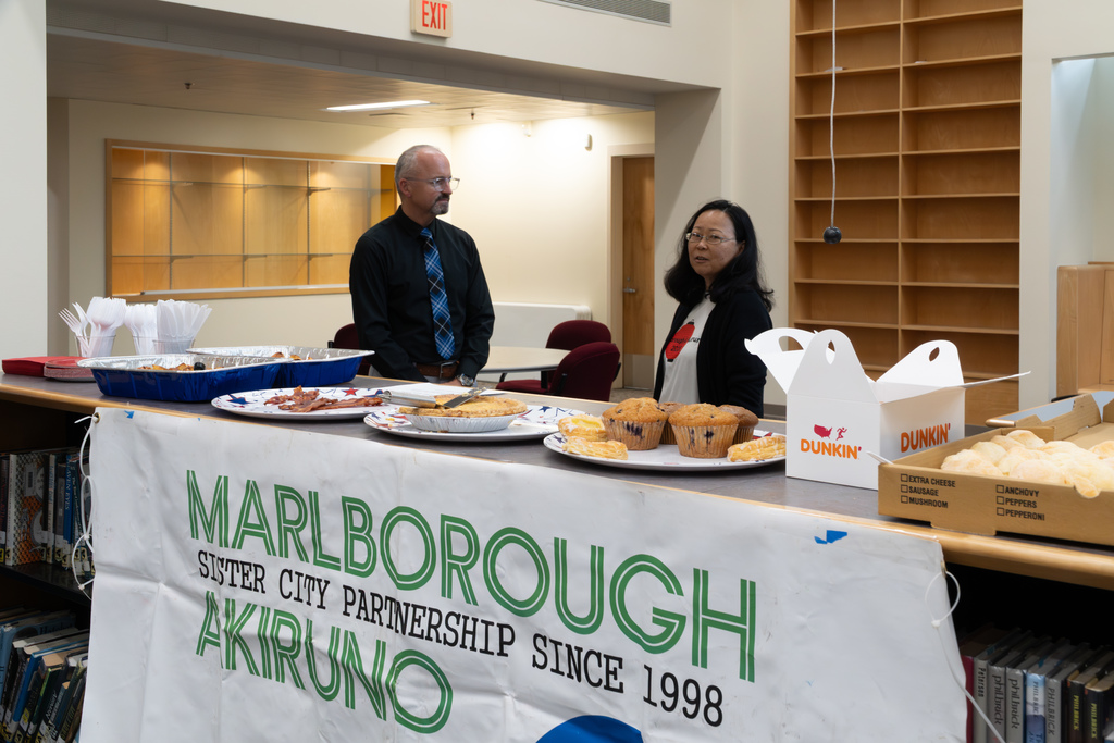 Interim Superintendent Jason Defalco and Ayako Barnum, a parent volunteer, stand and chat behind a table set with various breakfast items. A banner that reads "Marlborough-Akiruno Sister City Partnership" is displayed in front.