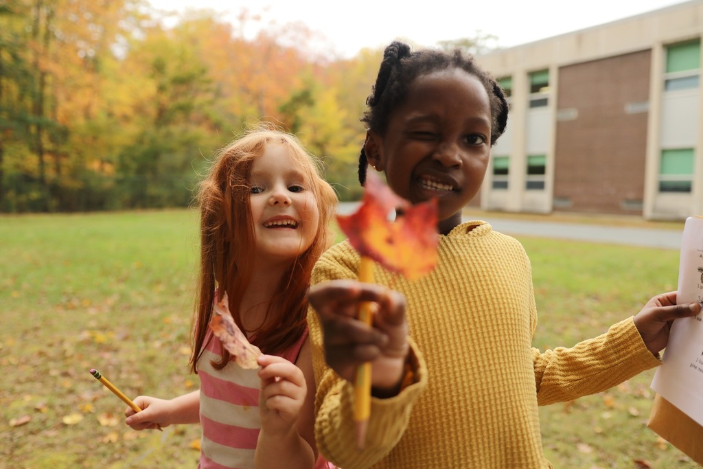 A young girl wearing a yellow pullover stands outside a school building holding a leaf and a pencil, winking at the camera. A young red-headed girl stands and smiles next to her.