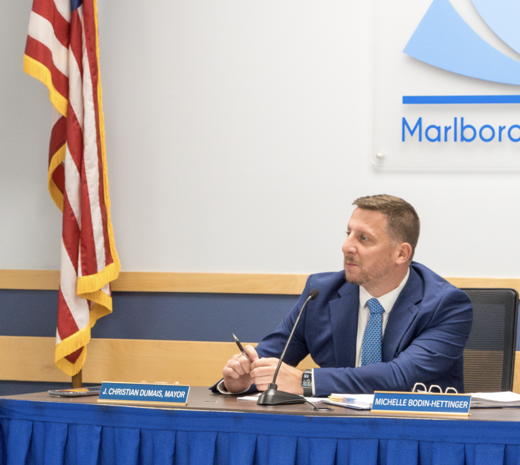 Marlborough School Committee Chair, Mayor Christian, sits at a table with the USA flag to his right and the MPS logo behind him. 