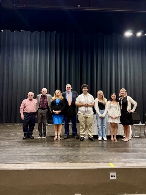 Three National Business Honor Society members pose for a group photo with school staff and committee members on stage in the Whitcomb Auditorium.