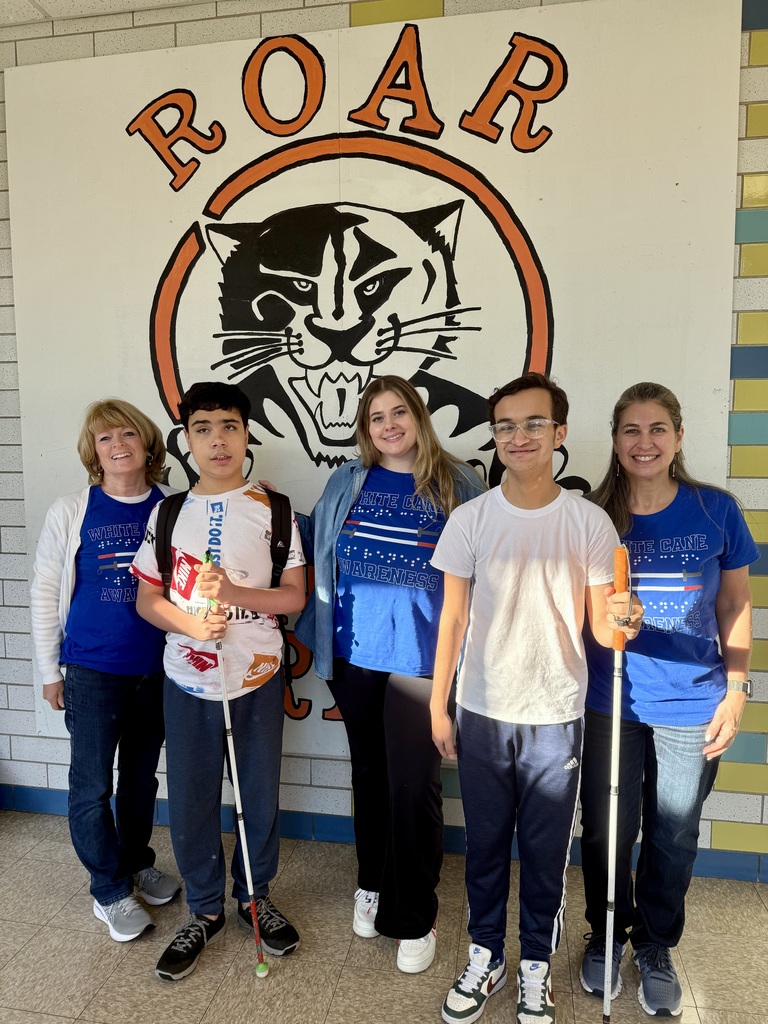 Two students who use white canes and three staff members pose for a group photo in front of the "Roar" mural at the Whitcomb School.