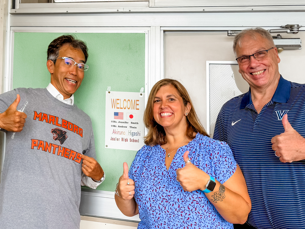 Two Whitcomb staff members and a Japanese man wearing a Marlboro Panthers t-shirt pose for a group photo in front of a welcome sign. All three are doing a double thumbs-up signal.