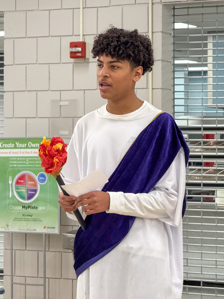 A student wearing a Roman robe costume holds a paper flower bouquet and a notecard.