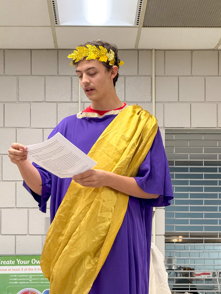 A student wearing a Romab robe costume and crown reads a script.
