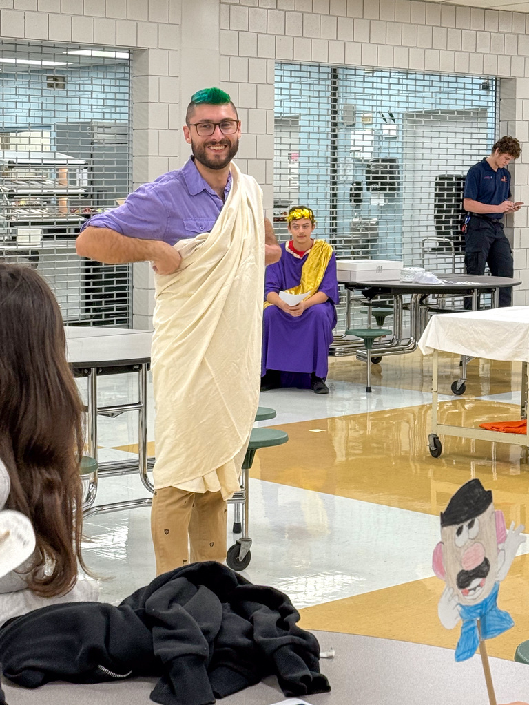 A smiling MHS teacher with green hair and glasses wears a Roman robe.