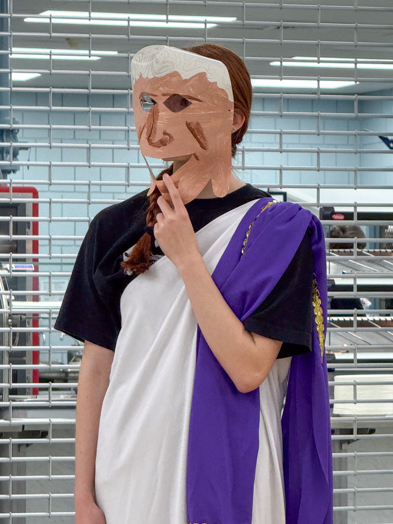 A student wearing a Roman robe costume and an imagine, a mask.