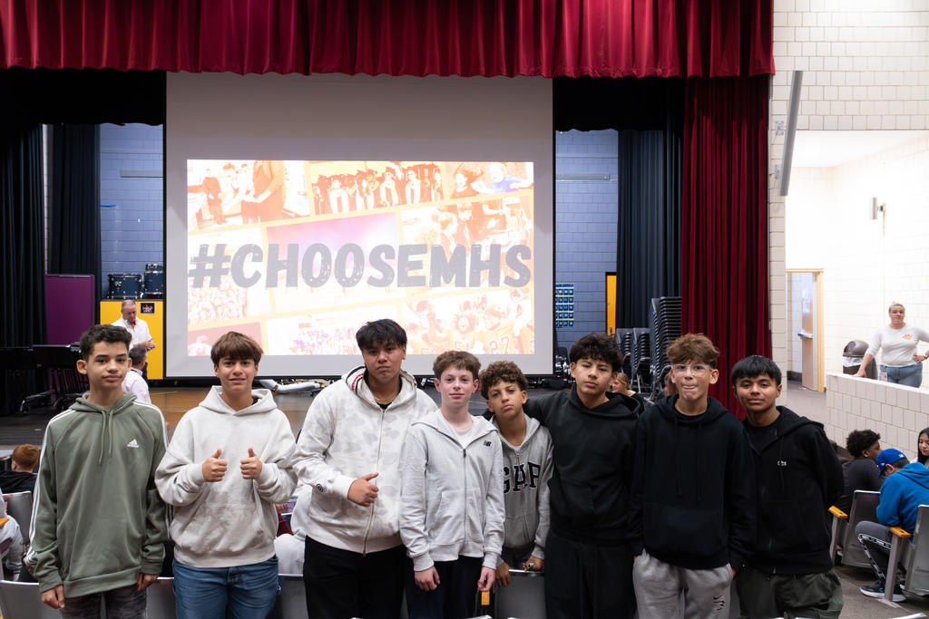 A group of 8th-grade boys pose for a group photo in front of a projector screen displaying a #ChooseMHS graphic.