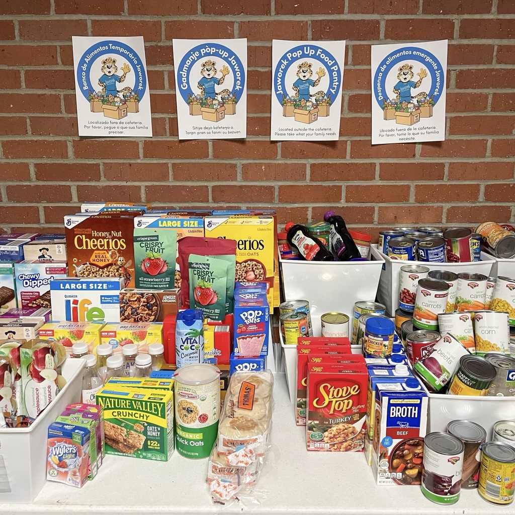 A table full of nonperishable items and canned goods. Four Jaworek flyers are posted on the brick wall behind the table.