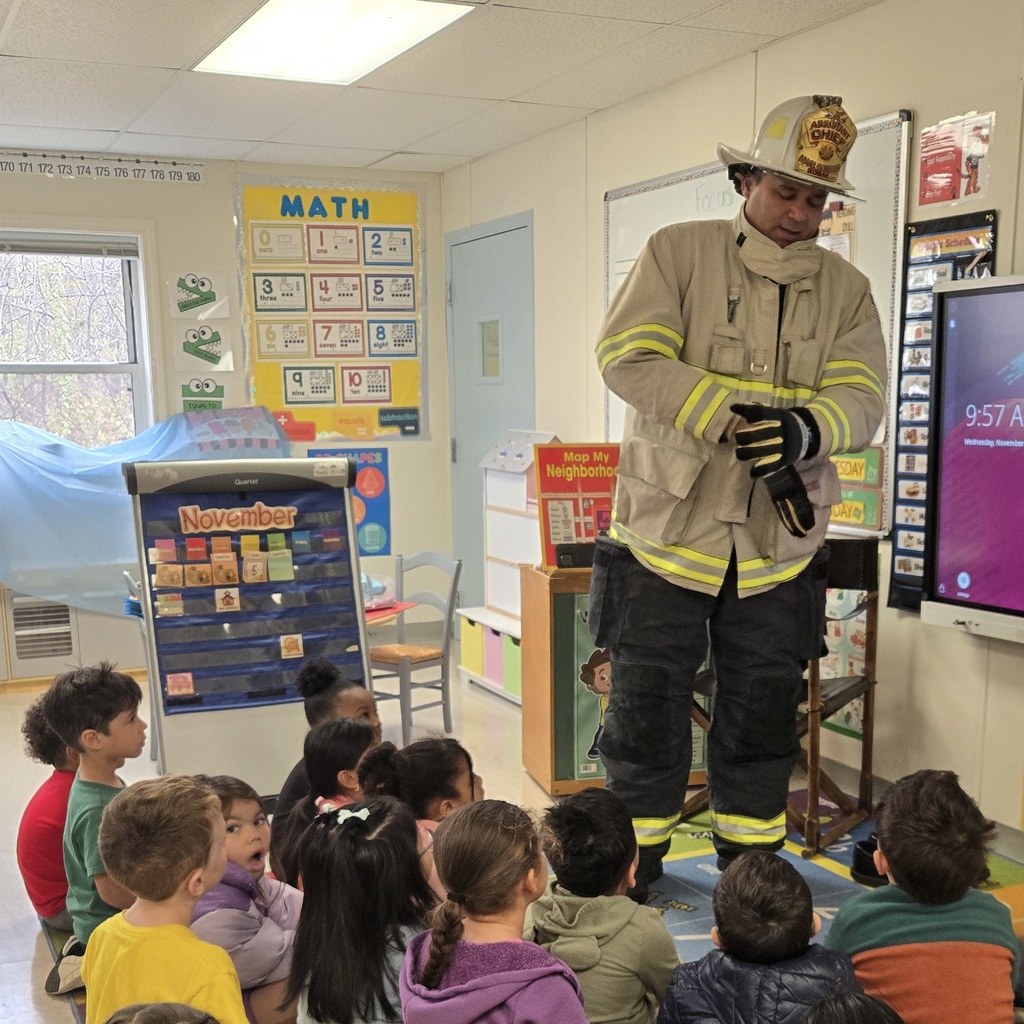 A firefighter stands and speaks to a group of Richer School students sitting on a rug.