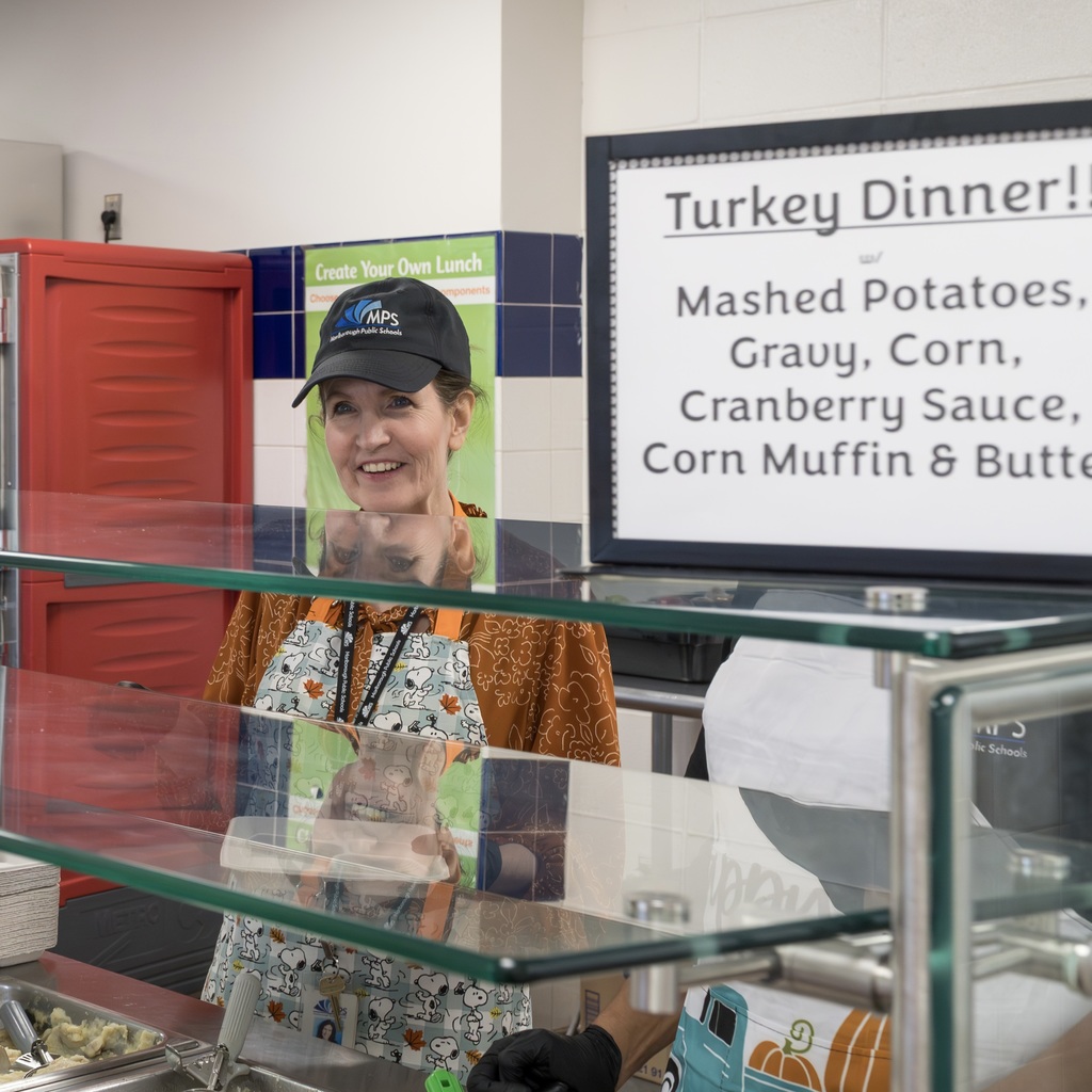 MPS Human Resources Director Patricia Brown stands behind the counter, smiling. A sign reads "Turkey Dinner! Mashed potatoes, gravy, corn, cranberry sauce, corn muffin & butter."
