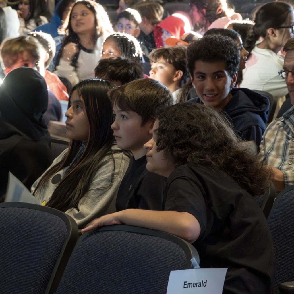  Three sixth-grade students sit in a row, looking into the distance. Another student sitting behind them smiles directly at the camera.