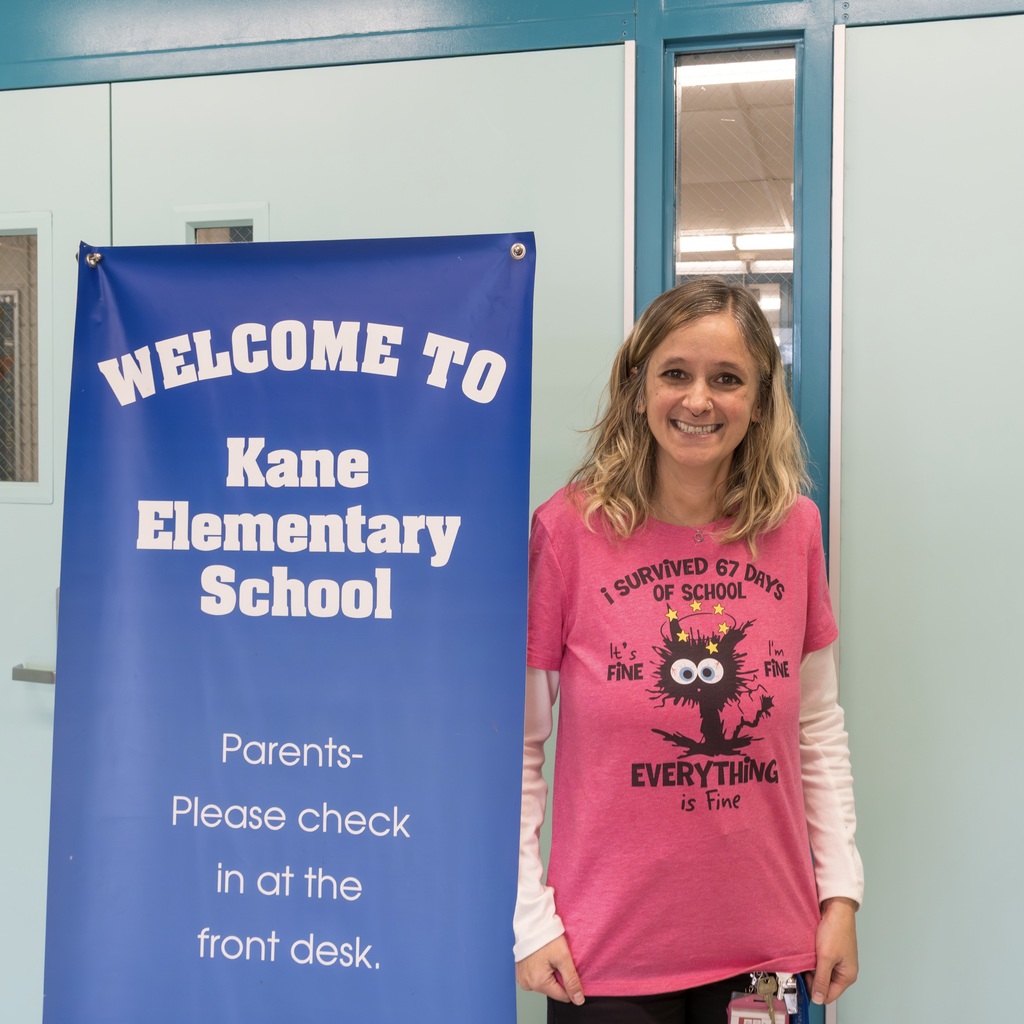 Kane School Principal stands next to a "Welcome to Kane Elementary School" banner, wearing a pink shirt that reads "I survived 67 days of school. It's fine. I'm fine. Everything is fine." with a drawing of a black cat.