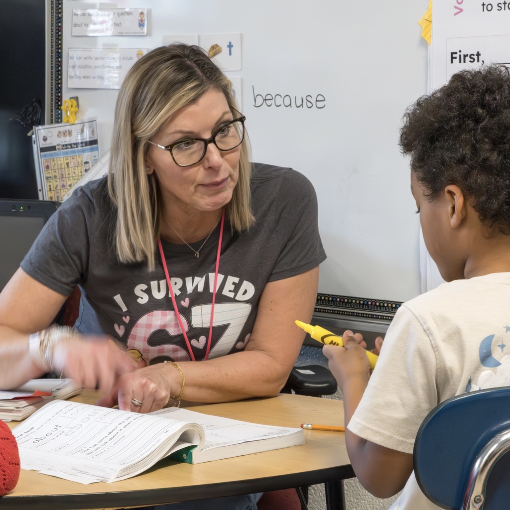 A Kane School teacher wearing a "I survived 67 days of school" shirt sits down with a student to complete a lesson.