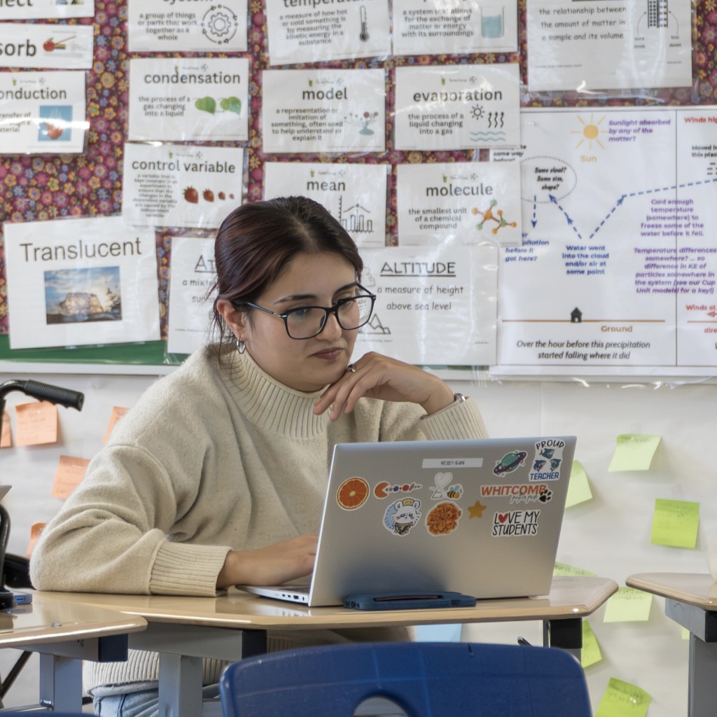 A teacher sits at their laptop in a classroom.