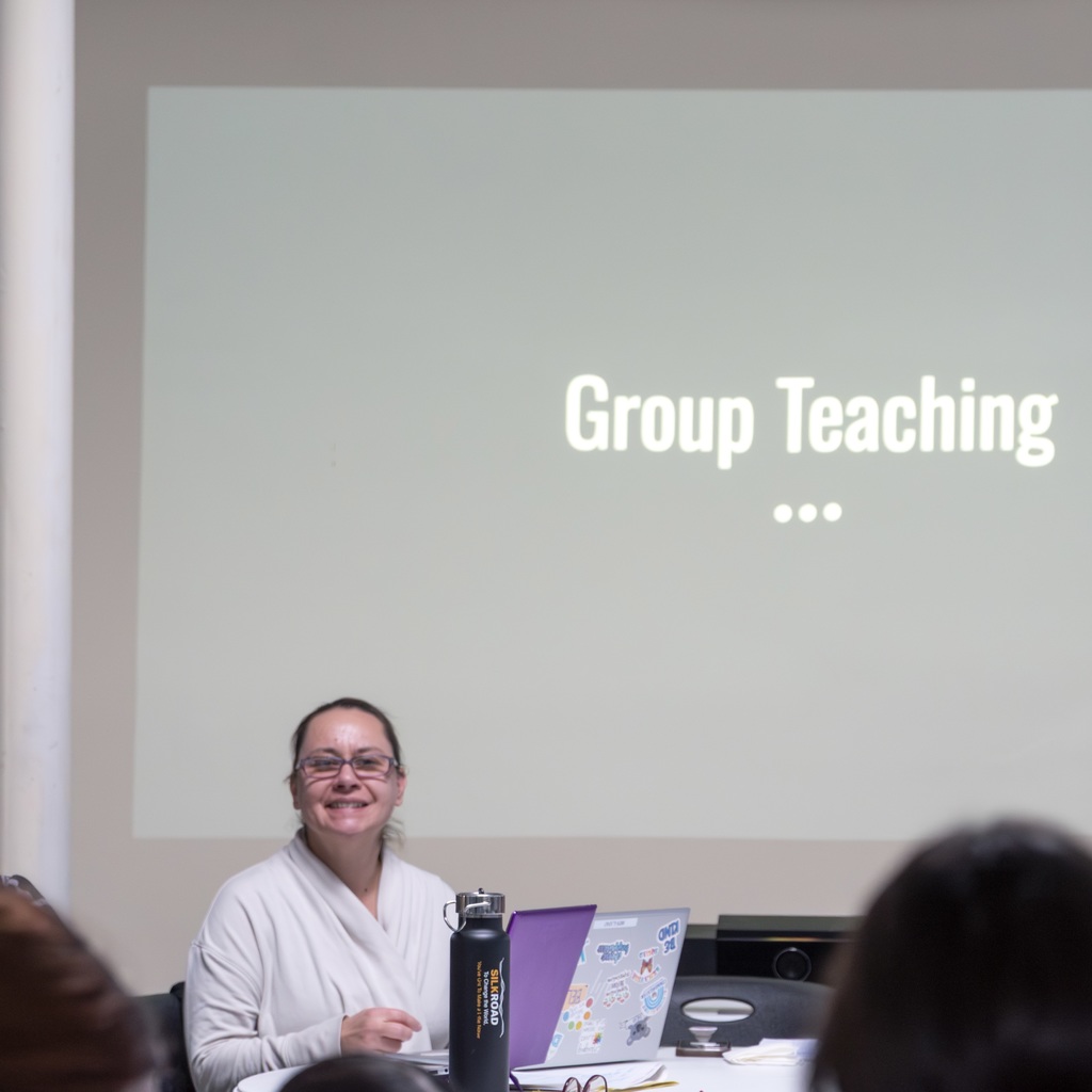 A teacher sits at a table. A slide reading "Group Teaching" is projected on the wall.