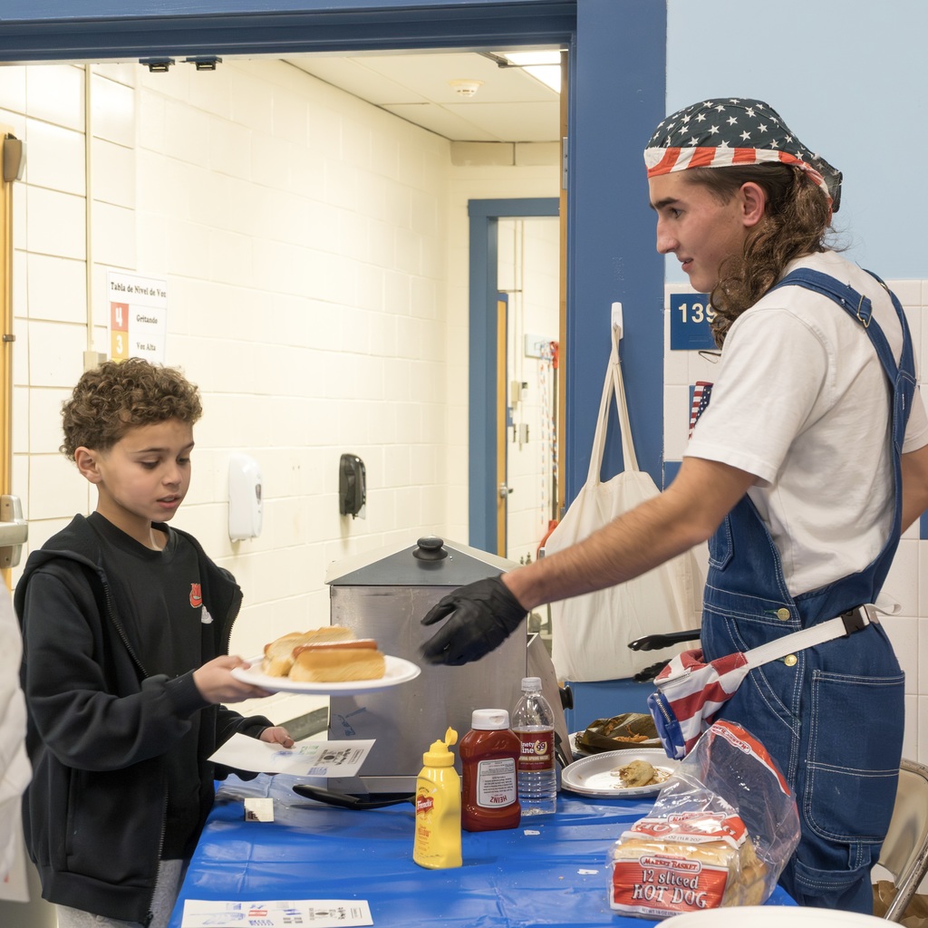 A Richer School staff member representing the USA hands out hot dogs to a student.