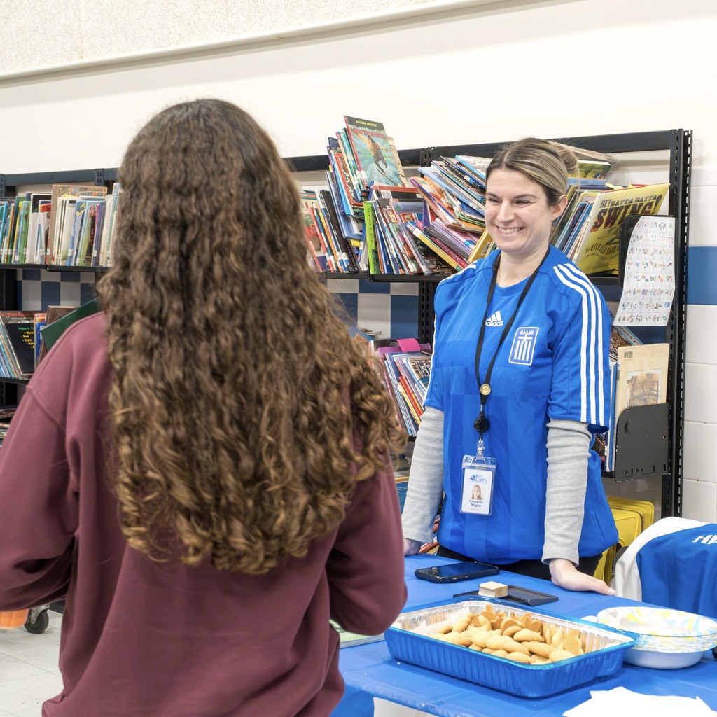 A Richer School representative from Greece stands behind a table with Greek food and smiles while talking to a student.