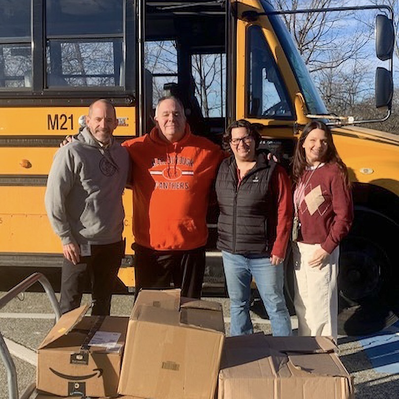 Two Richer School staff member and two NRT employees pose for a group photo in front of a yellow school bus. Three cardboard boxes with donations are in front of them.