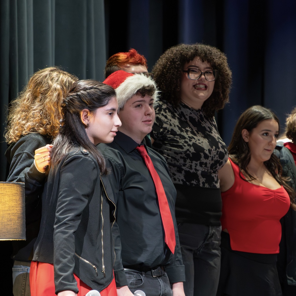 Voices ReChoired members and alums stand on stage, linking arms and hugging.