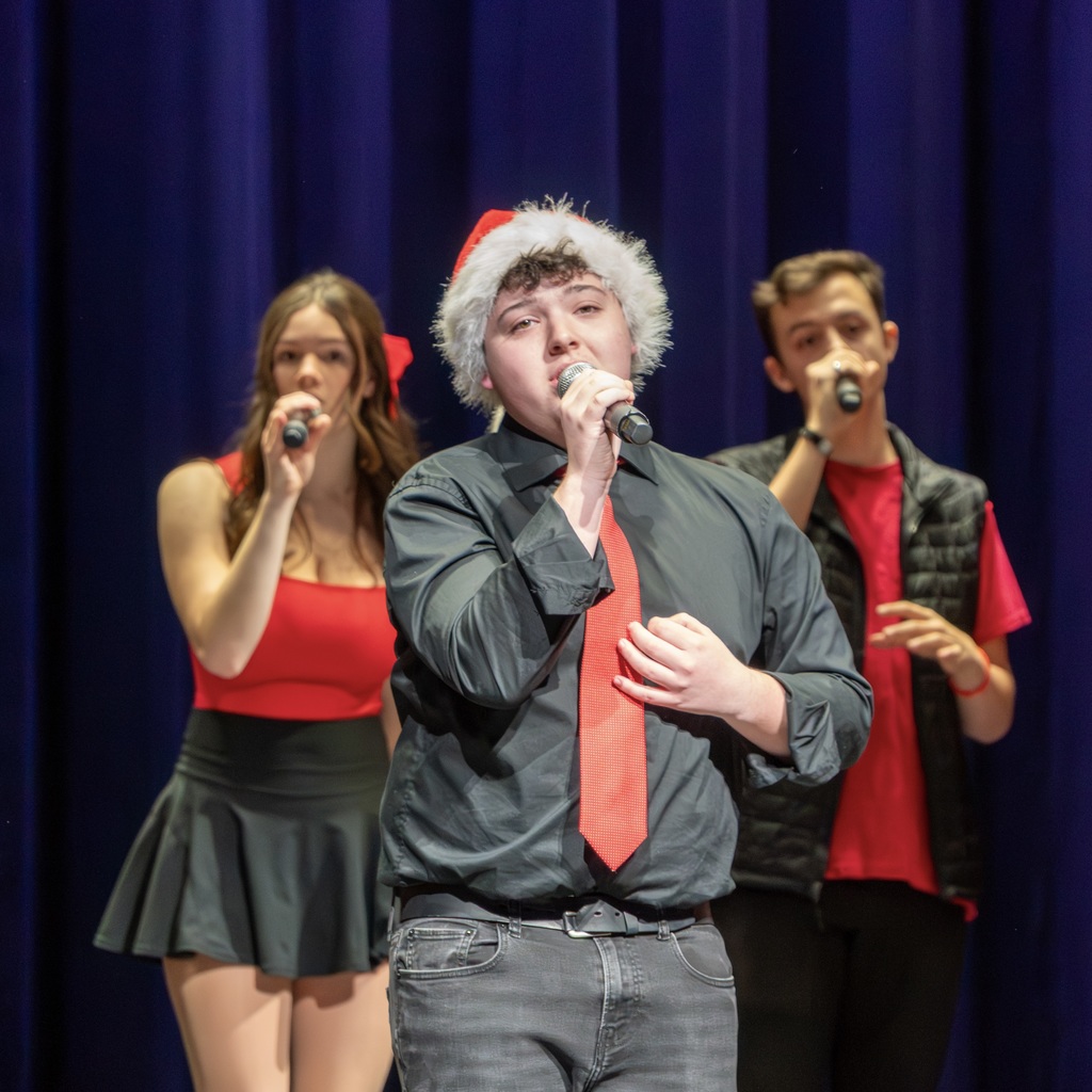 A Voices Rechoired member wearing a Santa hat sings a solo on stage. Two other members stand singing behind him.