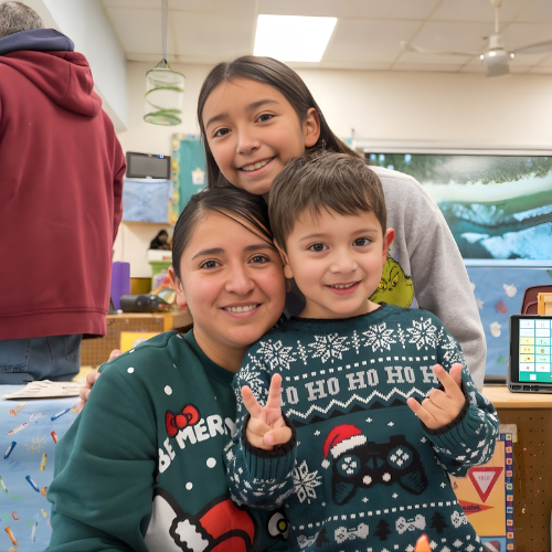 A preschool student, his mom, and his sister pose for a group photo, dressed in holiday sweaters.