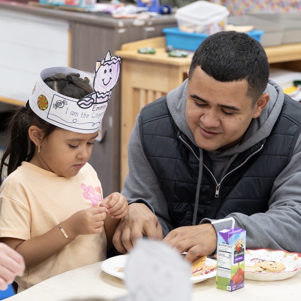 A smiling dad sits down at the table with his daughter to help her with a craft. The daughter is wearing a paper crown.