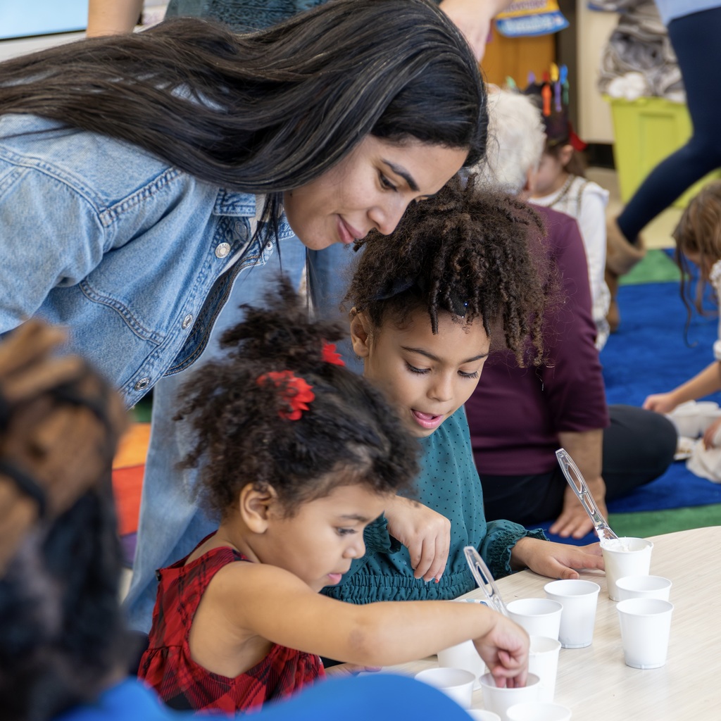 A mom smiles and stands over her two daughters as they sit at a table working on a craft.