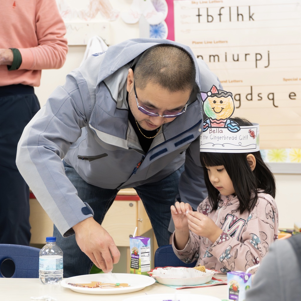 A dad stands over his daughter, who is sitting at a table, to help her with a craft.