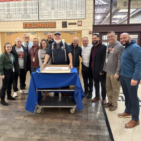 Jimmy O'Coin stands behind a cart with a cake. Several MHS and district employees stand with Jimmy.