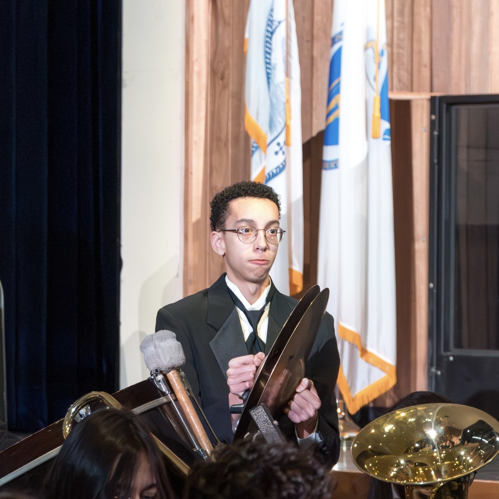 A Marlborough High School band member wearing a suit stands, playing the cymbal.