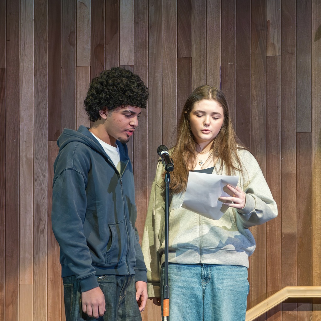 Two Whitcomb School students stand on stage and read the Pledge of Allegiance from a sheet of paper.