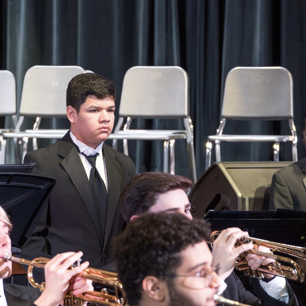 A Marlborough High School band student wearing a suit stands behind other band members sitting down, playing instruments.