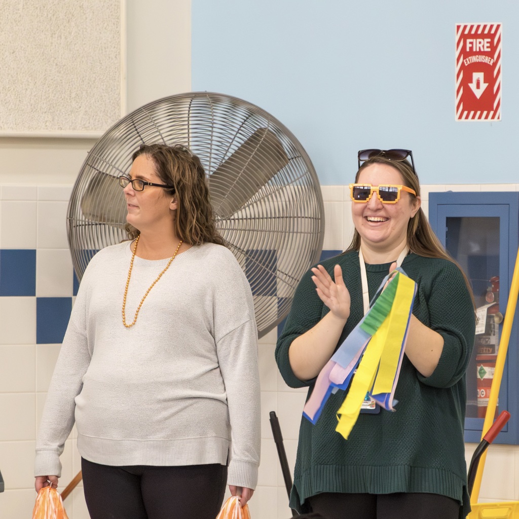Two Richer Schools staff members stand near a wall in the cafeteria. One staff member wearing glasses claps while holding a ribbon.