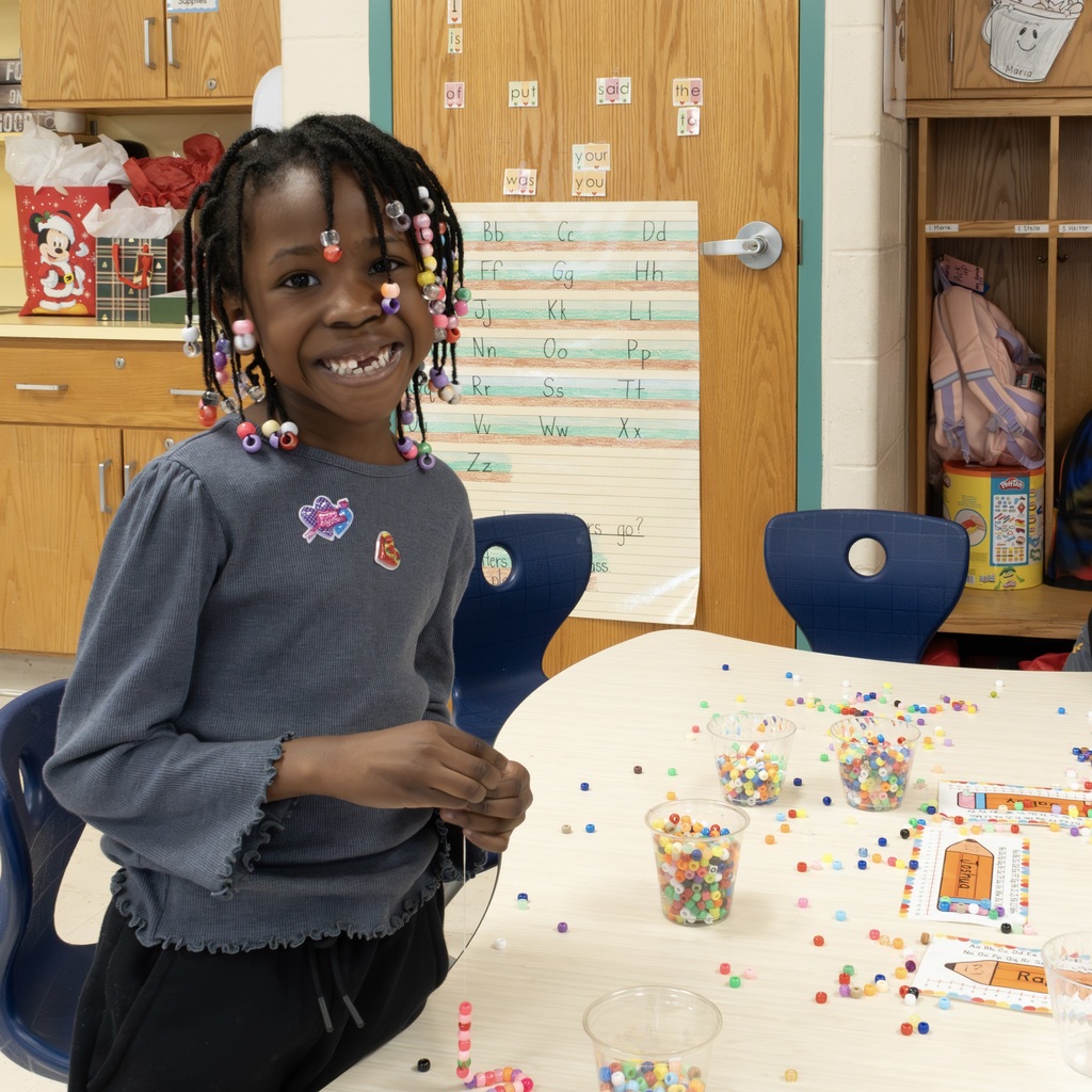 A student playing with beads stands and smiles at the camera.