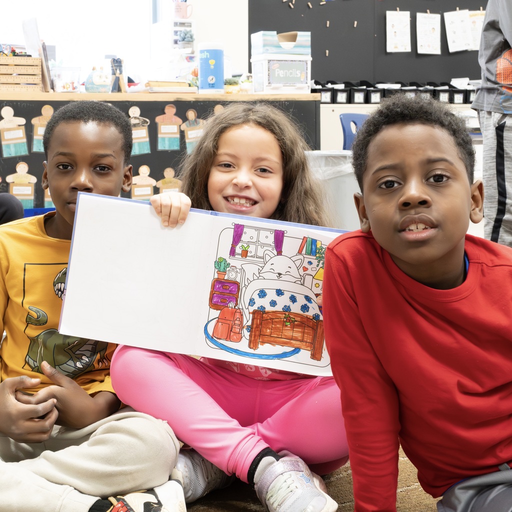 Three Jaworek students pose for a photo, sitting down on a rug. The girl in the middle holds up an open coloring book.