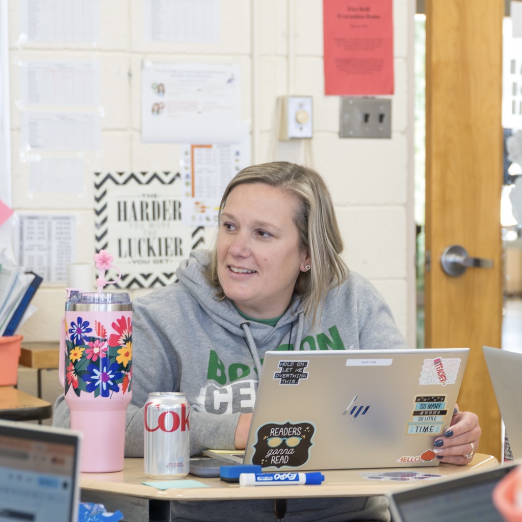 A female faculty member, sitting at a desk with an open laptop in front of her, smiles at someone out of the frame.