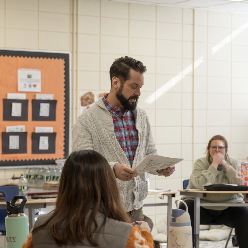MPS Assistant Superintendent RJ Skaza stands and reads from a sheet of paper in the middle of a room. Staff sit in desks in a circle around him.
