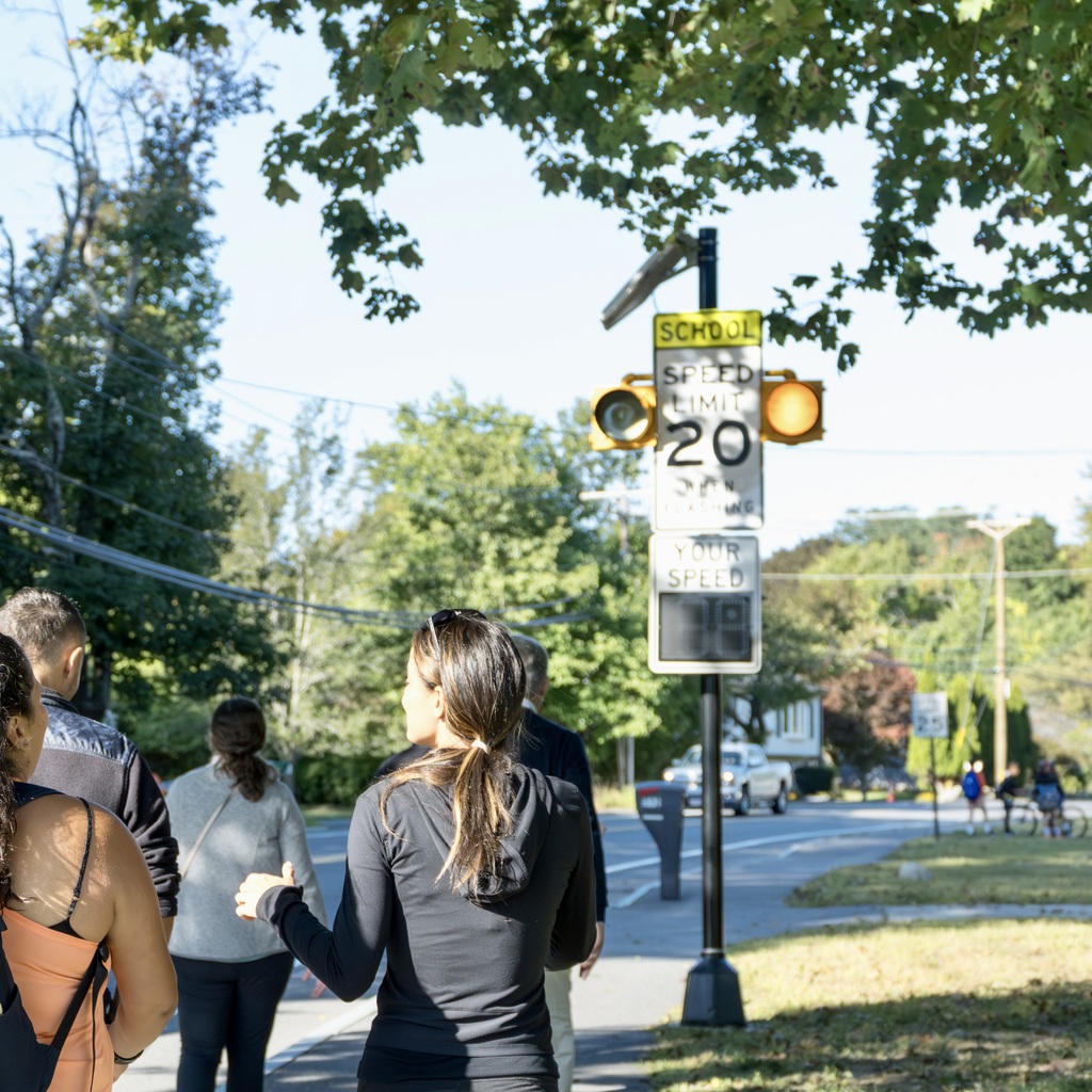A group of parents walk on a sidewalk behind a group of students biking and walking to school. A road sign with text that says 'SCHOOL SPEED LIMIT 20' is in front of them.