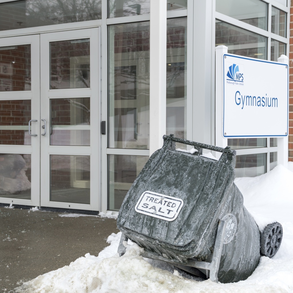 A treated salt bin is posted outside a building entrance. A sign behind the bin reads "Gymnasium."
