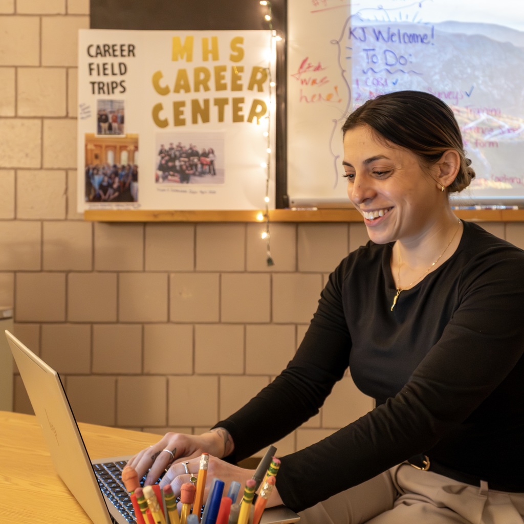 MHS Career Specialist Jackie Coyle smiles as she sits working on her laptop at a table. A poster reads "MHS Career Center" behind her.